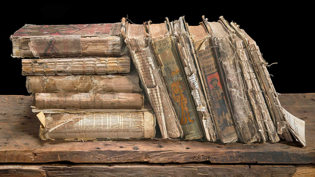 A detailed fine art still life of a leaning stack of distressed antique books on weathered wood against a black background.