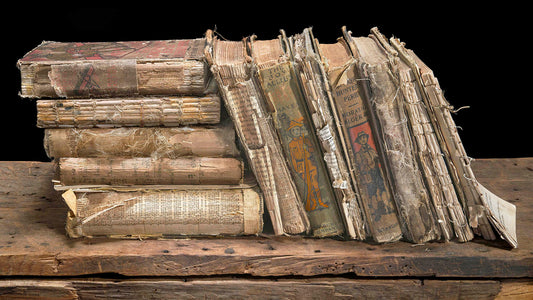 A detailed fine art still life of a leaning stack of distressed antique books on weathered wood against a black background.