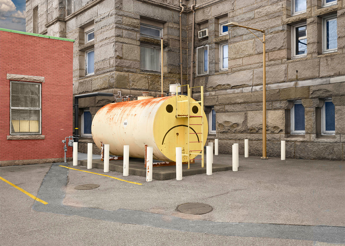 An environmental urban photography print of an old yellow oil storage tank named Fred with a painted frowning face tucked in a back alley.